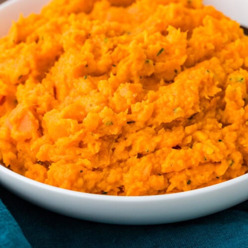 A close-up of a white bowl filled with creamy rosemary sweet potato mash. The vibrant orange dish rests on a teal fabric surface, hinting at a flavorful, comforting meal.