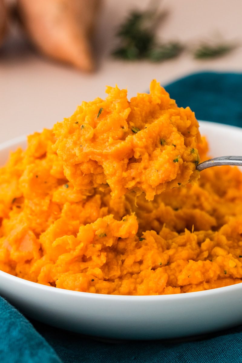 A spoonful of bright orange rosemary sweet potato mash with herbs held over a white bowl. The potatoes appear creamy and textured, with flecks of green herbs. A blue cloth is partially visible under the bowl.