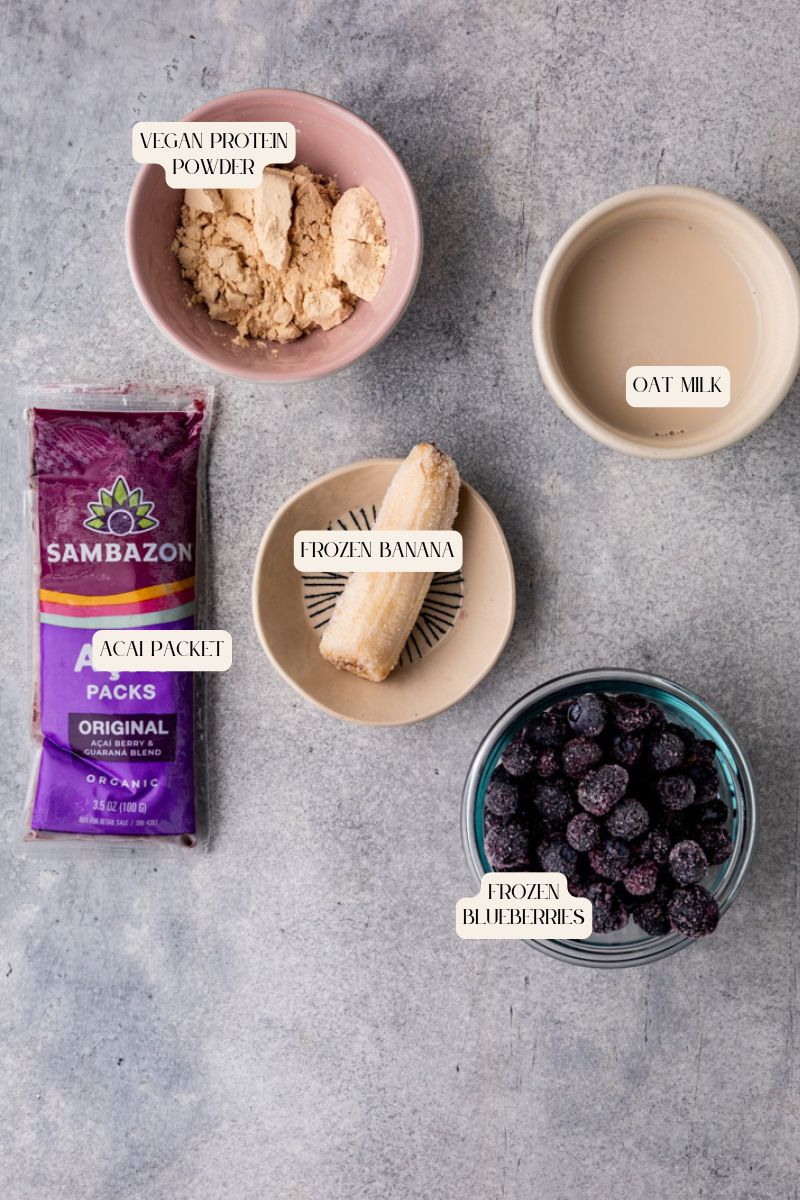 Ingredients for a smoothie arranged on a gray surface: a bowl of vegan protein powder, a dish of oat milk, a small bowl with a frozen banana, a package labeled Sambazon Açaí Packet, and a bowl of frozen blueberries.