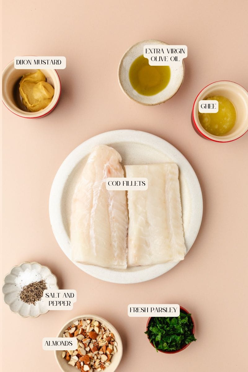 Top-down view of ingredients for an almond-crusted cod recipe arranged on a beige surface: cod fillets on a plate, small bowls of extra virgin olive oil, ghee, Dijon mustard, salt and pepper, chopped almonds, and fresh parsley.