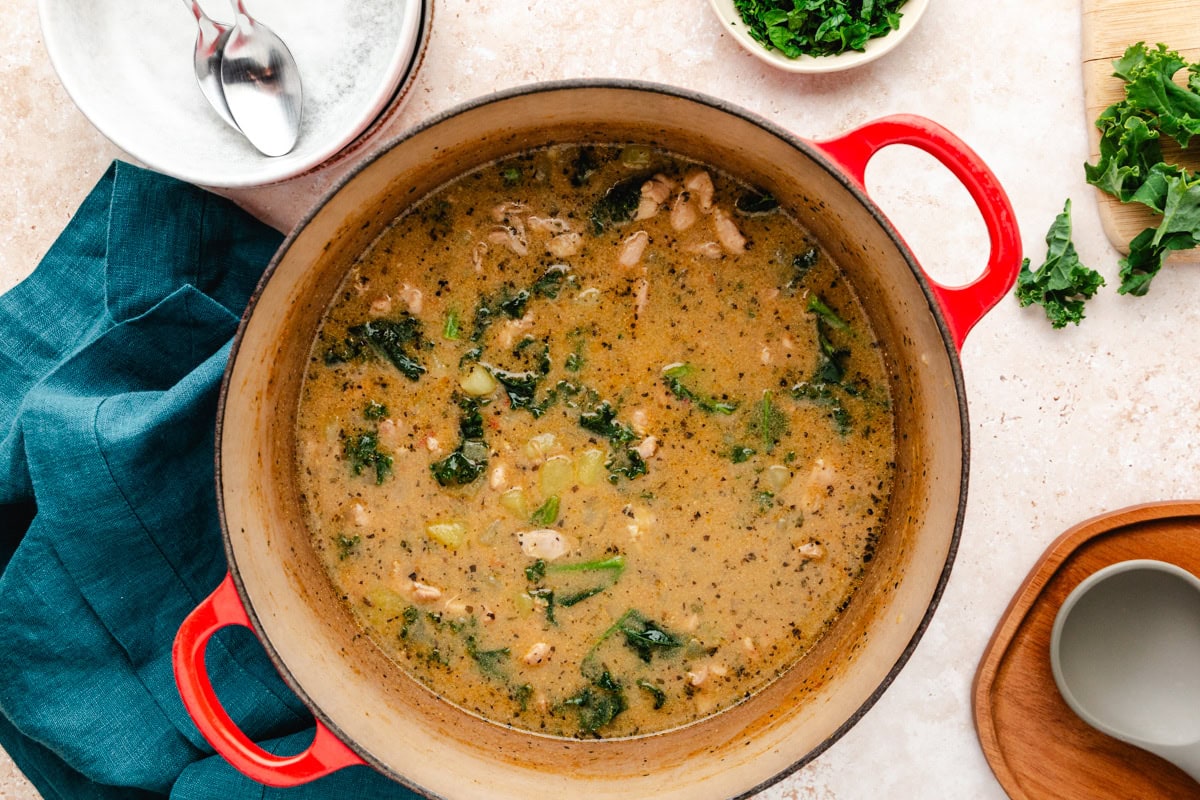 A red pot holds a hearty chicken and potato soup with chunks of meat, leafy greens, and a creamy broth. Spoons rest on a saucer nearby, and a bowl of chopped greens is positioned beside a teal napkin. The setting suggests cozy meal preparation with this comforting recipe.
