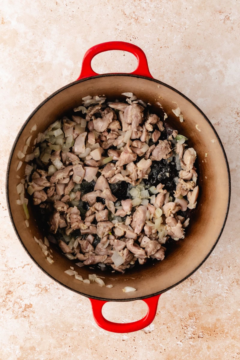 A red pot filled with chopped onions, potatoes, and pieces of chicken simmers on the stove. The ingredients for this chicken and potato soup recipe are lightly browned and mixed together, resting on a beige countertop.