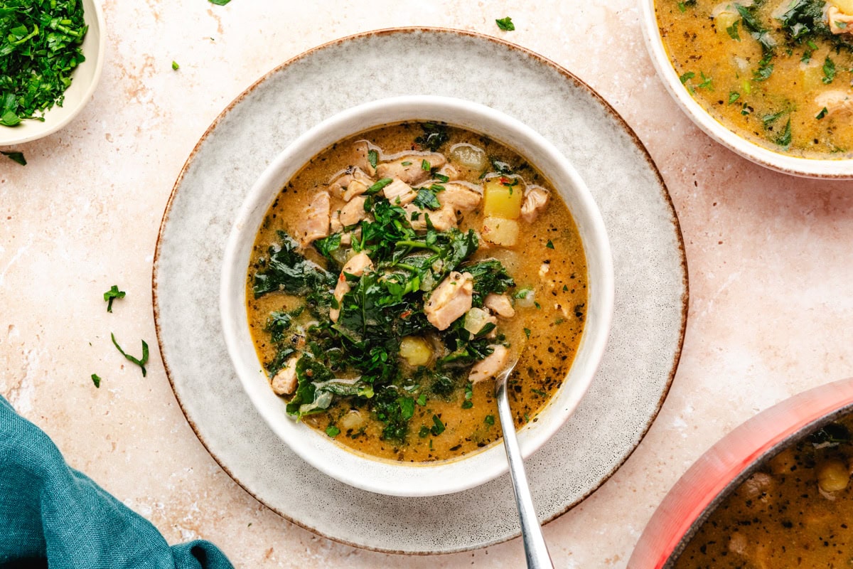 A bowl of hearty chicken and potato soup recipe is served on a speckled white dish. The soup is filled with chunks of chicken, leafy kale, and potato pieces, garnished with fresh parsley. A spoon rests inside the bowl, and a small bowl of chopped herbs is nearby.