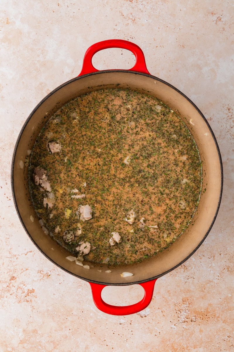 A red-handled pot contains a savory chicken and potato soup recipe with visible herbs and tender chicken pieces, set against a light, textured surface.