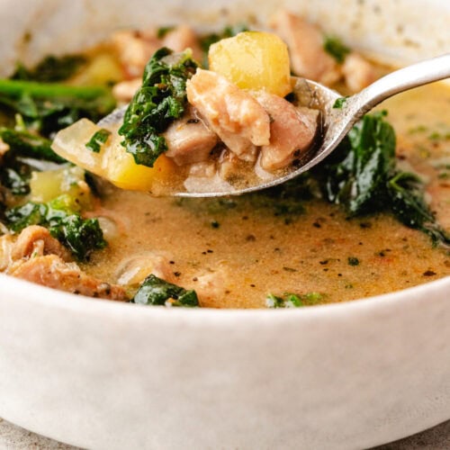 Close-up of a spoon lifting a delicious chicken and potato soup from a ceramic bowl. The creamy broth, dotted with chunks of meat, tender potatoes, and wilted greens, showcases a hearty texture and an inviting blend of green and beige colors.