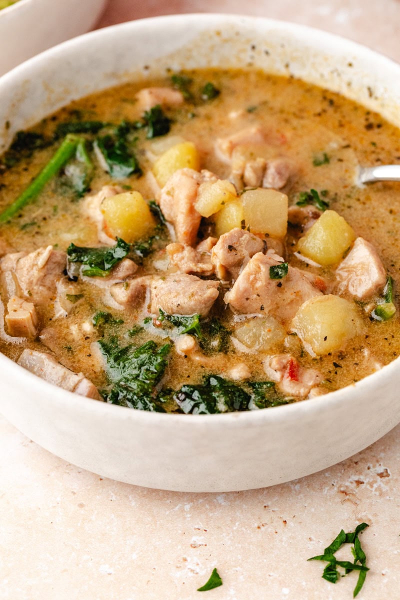 A bowl of hearty chicken and potato soup features chunks of meat, diced potatoes, and leafy greens in a savory broth. A spoon rests in the bowl, set on a light-colored surface. The dish appears warm and comforting—perfect for anyone seeking a soothing chicken and potato soup recipe.