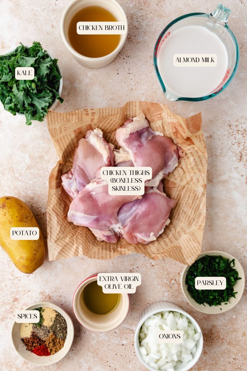 A top-down view of various ingredients for a chicken and potato soup recipe: raw boneless chicken thighs on paper, surrounded by bowls of kale, chicken broth, almond milk, chopped onions, parsley, spices, extra virgin olive oil, and a whole potato.