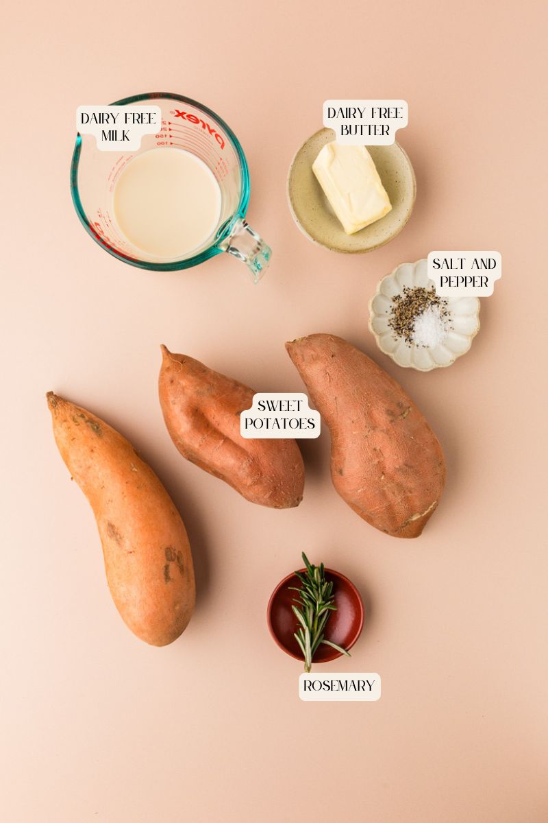 A flat lay on a beige surface features ingredients for rosemary sweet potato mash: sweet potatoes, dairy-free milk in a measuring cup, dairy-free butter on a plate, and salt and pepper in a dish. A small bowl of rosemary adds aromatic flair. Labels identify each item.