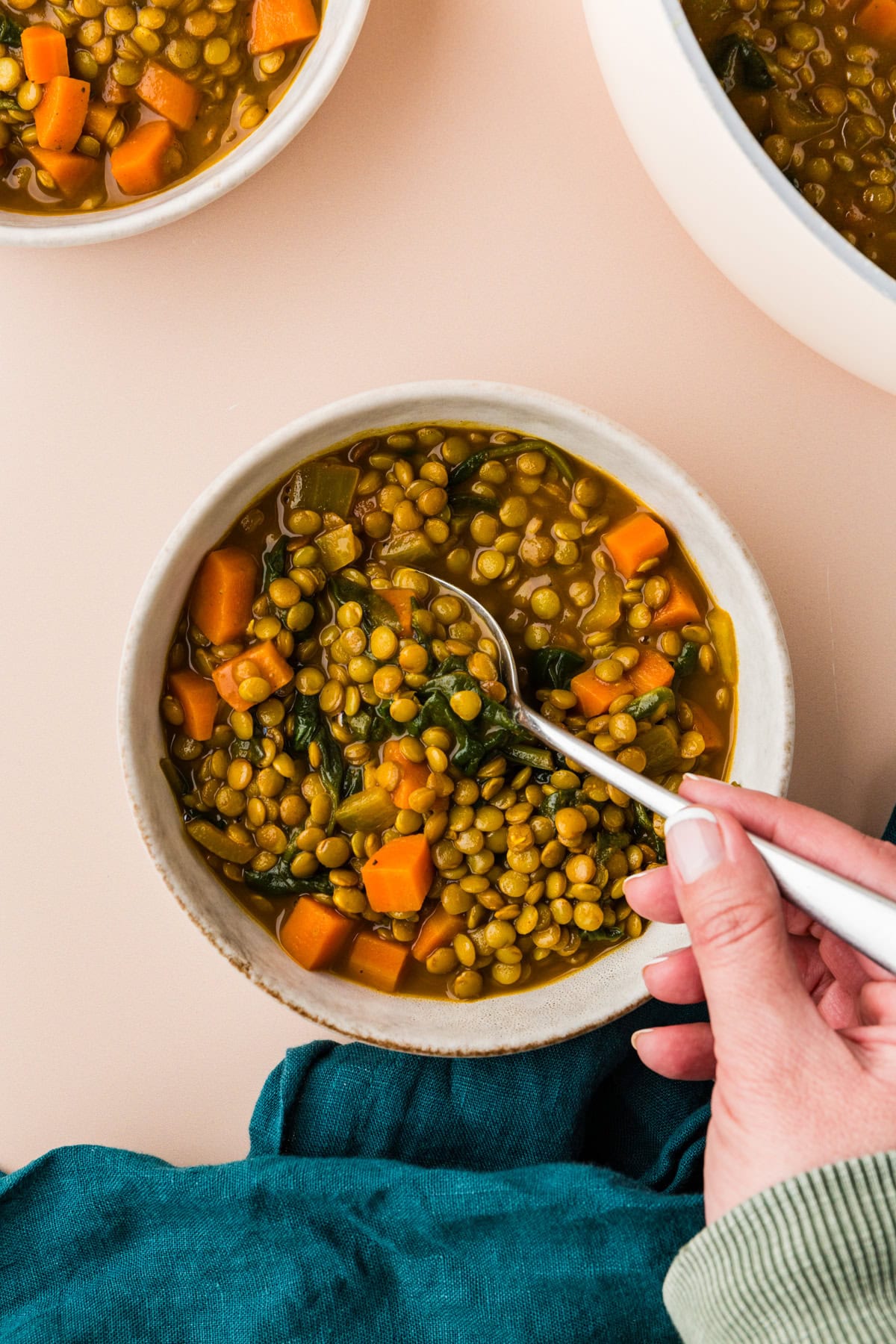 A hand gracefully holds a spoon over a bowl of carrot lentil soup, rich with green lentils, diced carrots, and fresh greens. The bowl rests on a light surface adorned with dark green fabric, while more soup tantalizingly peeks from the background.