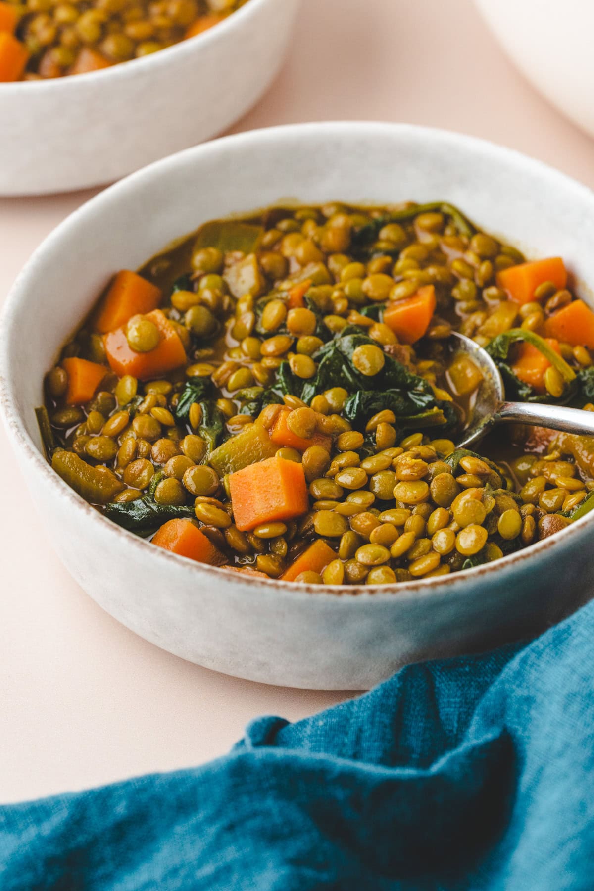 A bowl of carrot lentil soup with visible pieces of carrots and spinach. The soup appears rich and hearty, with a spoon resting in the bowl. A blue cloth napkin is draped on the surface nearby.