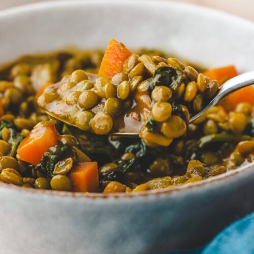 Close-up of a bowl of carrot lentil soup. The spoon holds a scoop with lentils, carrots, and greens. The blurred background hints at another dish and a blue cloth.