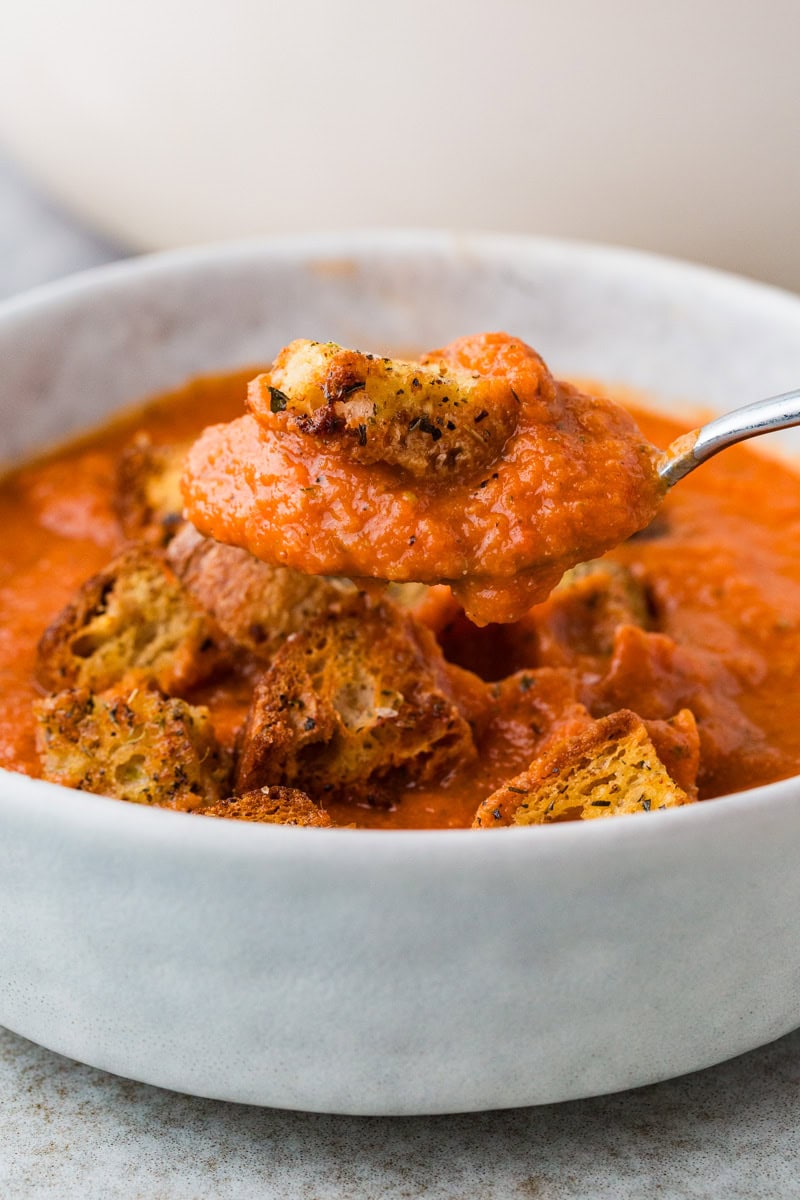 A bowl of creamy heirloom tomato soup topped with croutons. A spoon lifts a serving, showcasing the rich texture of the soup and the crispy, seasoned croutons. The dish is set in a light-colored bowl on a gray surface.