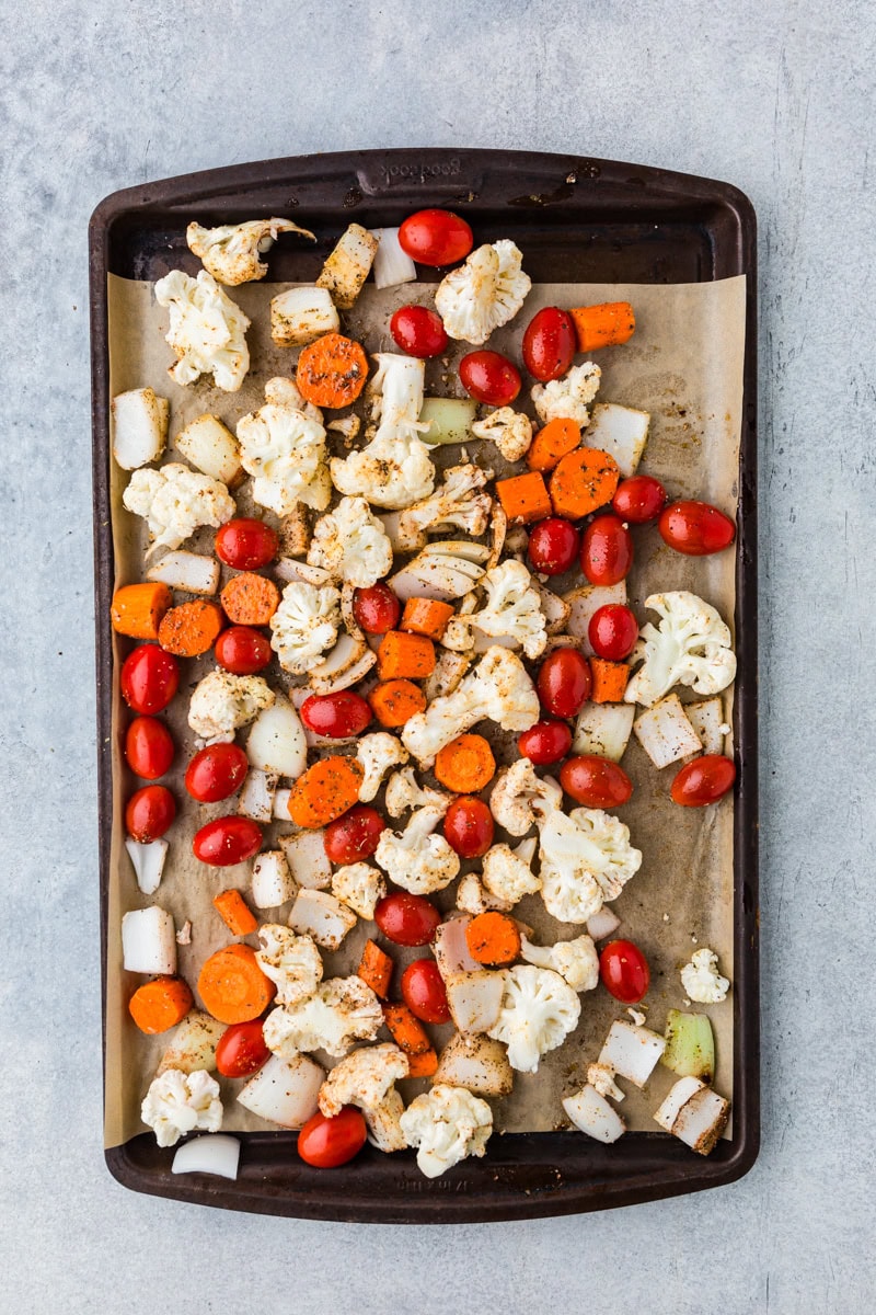 A baking tray with seasoned vegetables, including cauliflower florets, heirloom cherry tomatoes, sliced carrots, and chunks of onion, beautifully arranged on parchment paper and ready for roasting to create a delightful base for heirloom tomato soup.