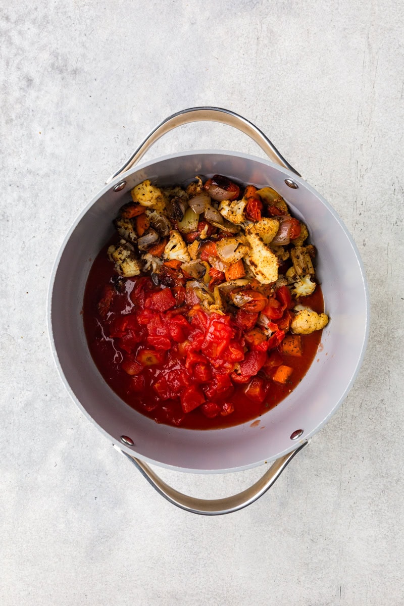 A pot containing heirloom tomato soup with chopped tomatoes and roasted vegetables like onions, garlic, and peppers. The pot’s silver handles are visible against a light gray background.