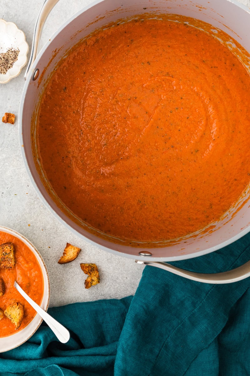 A large pot of heirloom tomato soup with herbs sits on a gray countertop. Beside it, a bowl is filled with the same soup and croutons. Croutons are scattered around, and a dark green cloth is draped across the table.