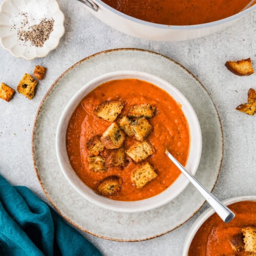 A bowl of heirloom tomato soup topped with croutons sits on a round plate beside a blue cloth. Another bowl is partially visible, along with a dish of black pepper. A pot of the flavorful soup is also visible in the background.