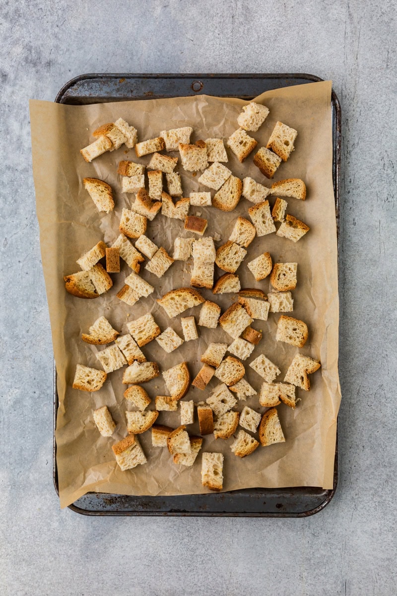 Cubed bread pieces spread evenly on a baking tray lined with parchment paper. The tray is on a textured gray surface.