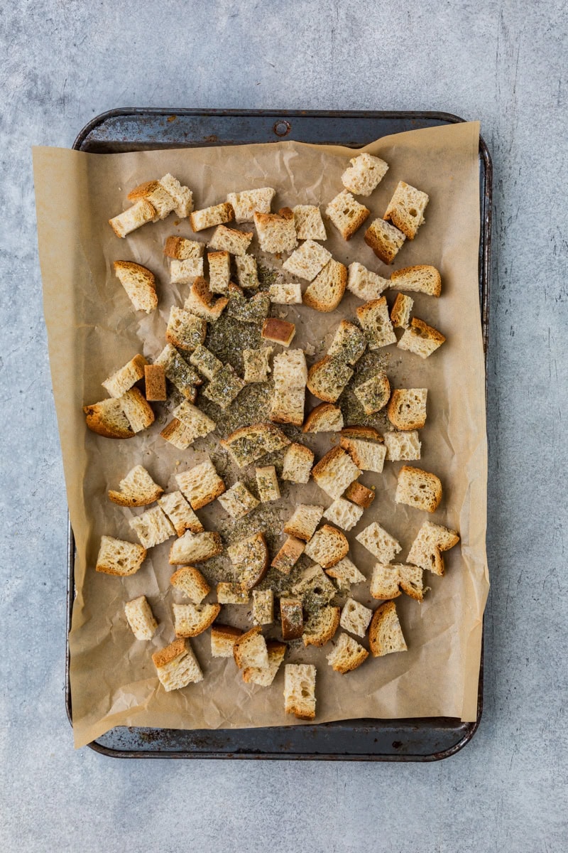 Chopped bread cubes seasoned with herbs are spread on a parchment-lined baking sheet.