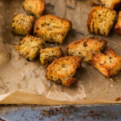Golden, seasoned croutons are scattered on a parchment-lined baking sheet. The croutons are crispy and varied in size, with visible herbs and a rustic texture, against a teal cloth on the side.