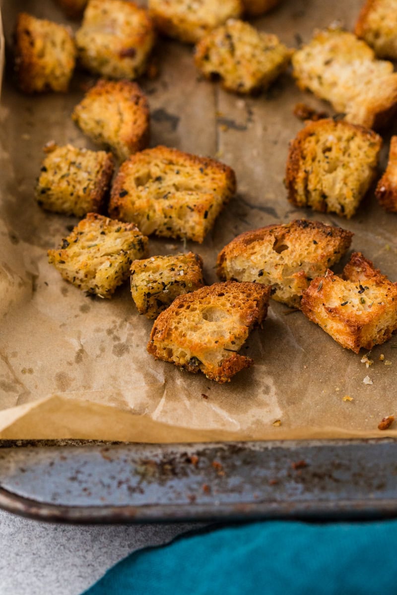 Golden, seasoned croutons are scattered on a parchment-lined baking sheet. The croutons are crispy and varied in size, with visible herbs and a rustic texture, against a teal cloth on the side.