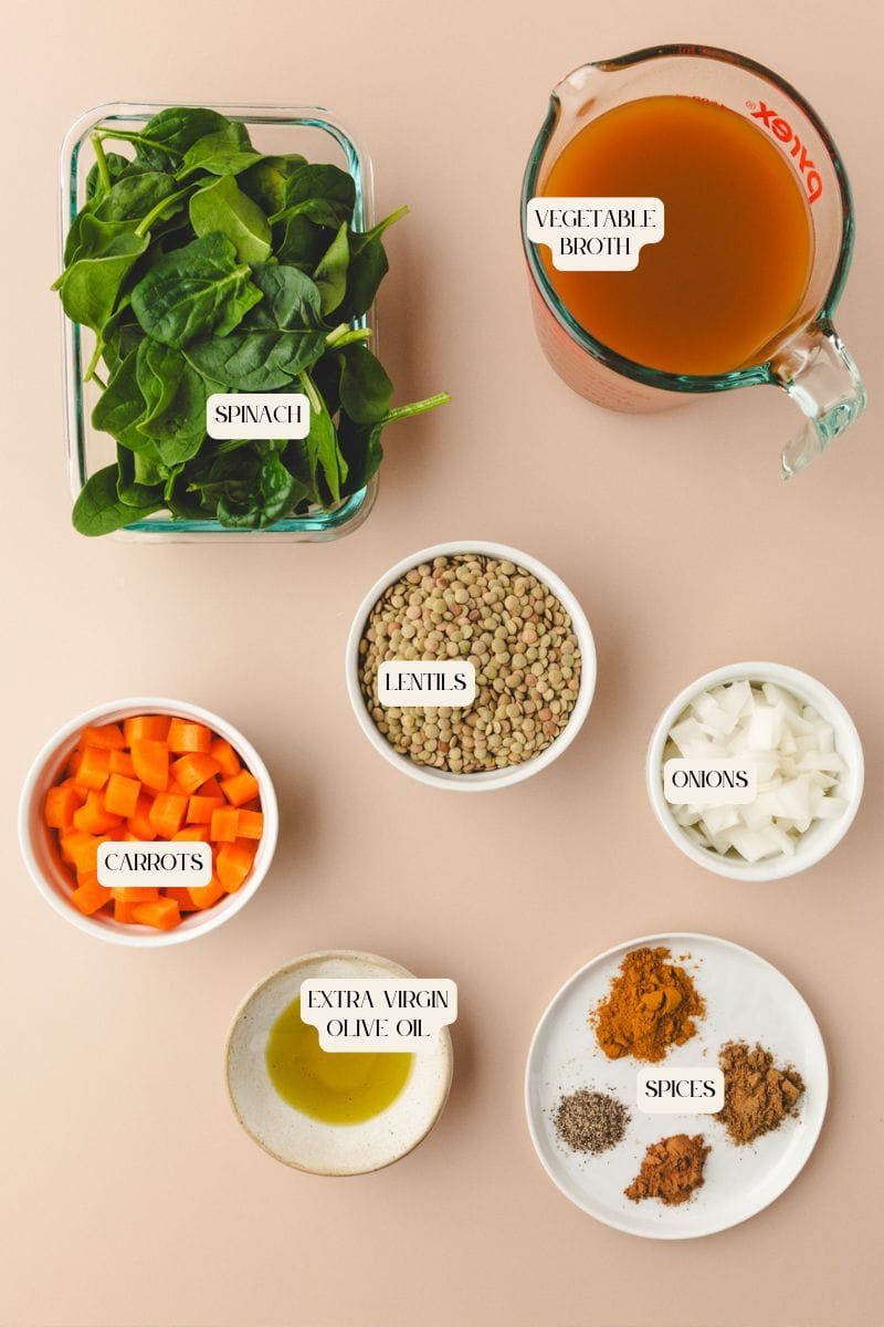 Ingredients for a carrot lentil soup are neatly arranged on a beige background: fresh spinach, vegetable broth in a measuring cup, dry lentils, chopped onions, extra virgin olive oil, diced carrots, and a plate of various spices.