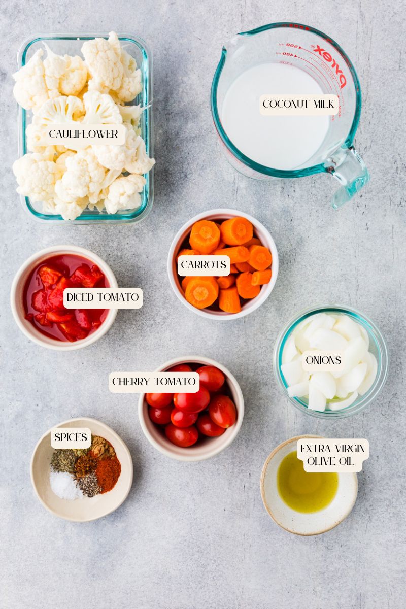 Top view of raw ingredients labeled and arranged on a gray surface for an heirloom tomato soup, including cauliflower, coconut milk, diced tomato, carrots, onions, cherry tomatoes, spices, and extra virgin olive oil.