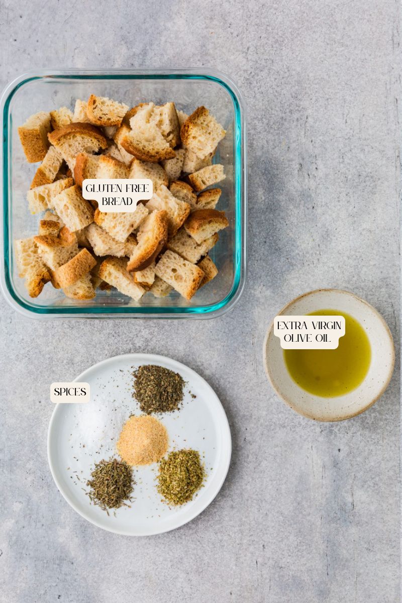 A glass container with cubed gluten-free croutons sits alongside a small bowl of extra virgin olive oil and a plate featuring spices like Italian herbs, garlic powder, and oregano. All items are artfully arranged on a gray surface.