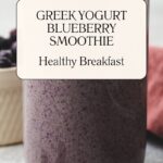 A glass brimming with a thick, purple concoction tagged Greek Yogurt Blueberry Smoothie and Healthy Breakfast stands proudly. In the backdrop, a bowl of berries adds charm. Discover more at whensaltyandsweetunite.com.