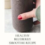 A hand with red nail polish holds a glass of blueberry smoothie blended with Greek yogurt, against a blurred background of blueberries and a cloth. Text on image: Healthy Blueberry Greek Yogurt Smoothie Recipe with a Healthy Breakfast badge.