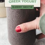 A hand holding a glass of creamy greek yogurt blueberry smoothie, with a green sweater sleeve visible. The text on the image reads, Healthy and Simple Greek Yogurt Blueberry Smoothie. A background of blurred objects adds context to the setting.