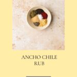 A bowl of various spices, neatly arranged on a light marble surface, showcases the rich flavors of an ancho rub. The text reads Ancho Chile Rub above Homemade Spice Blends. Visit whensaltyandsweetunite.com for more.