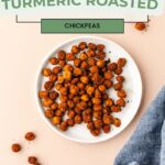 A white plate teeming with turmeric roasted chickpeas rests on a light beige surface, with a few chickpeas scattered around. A hint of blue cloth peeks from the right. Text reads: Healthy Snack Ideas - Turmeric Roasted Chickpeas.