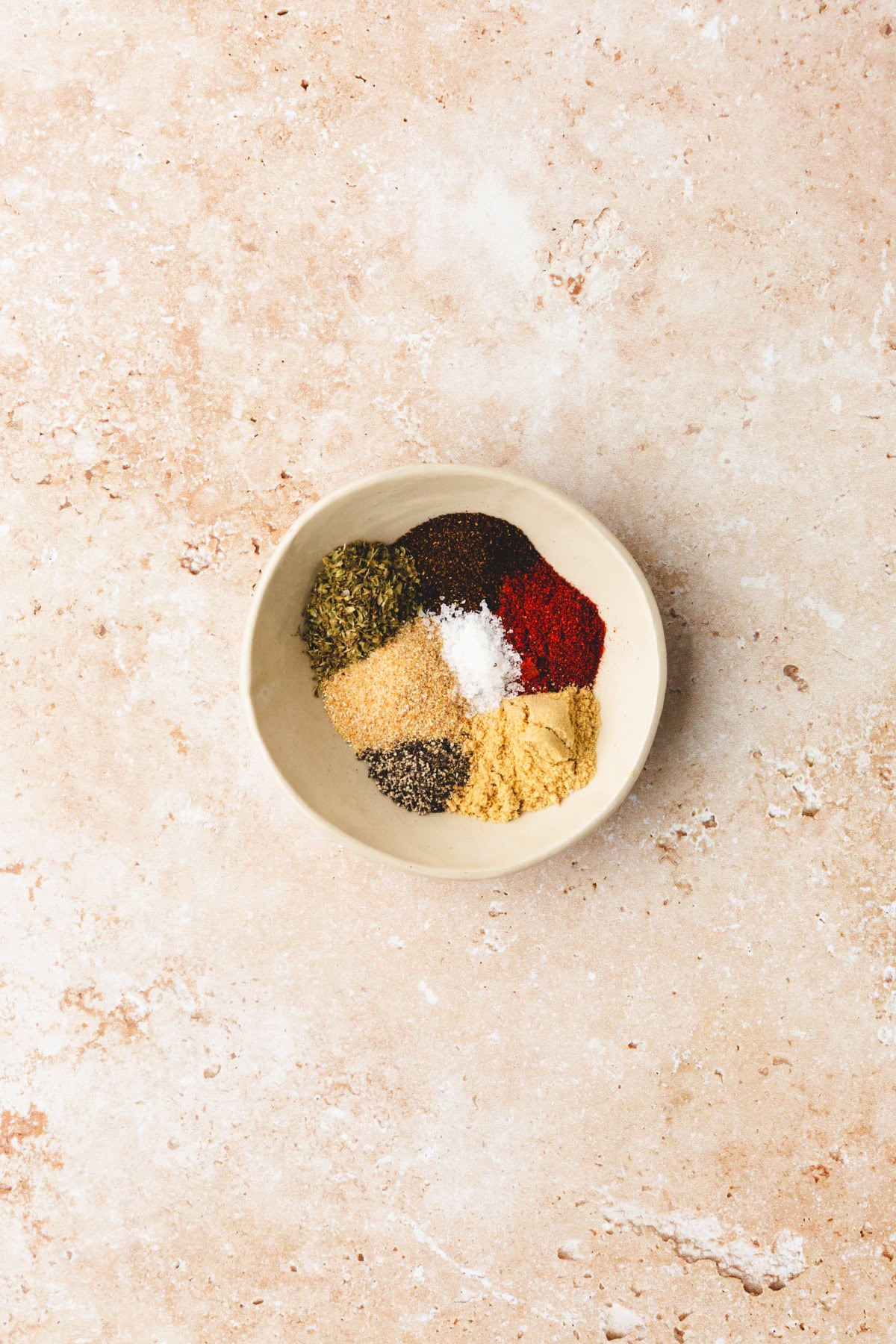 A bowl filled with various spices, each in separate small mounds.Among them lies an enticing ancho rub. The bowl rests on a textured beige surface.