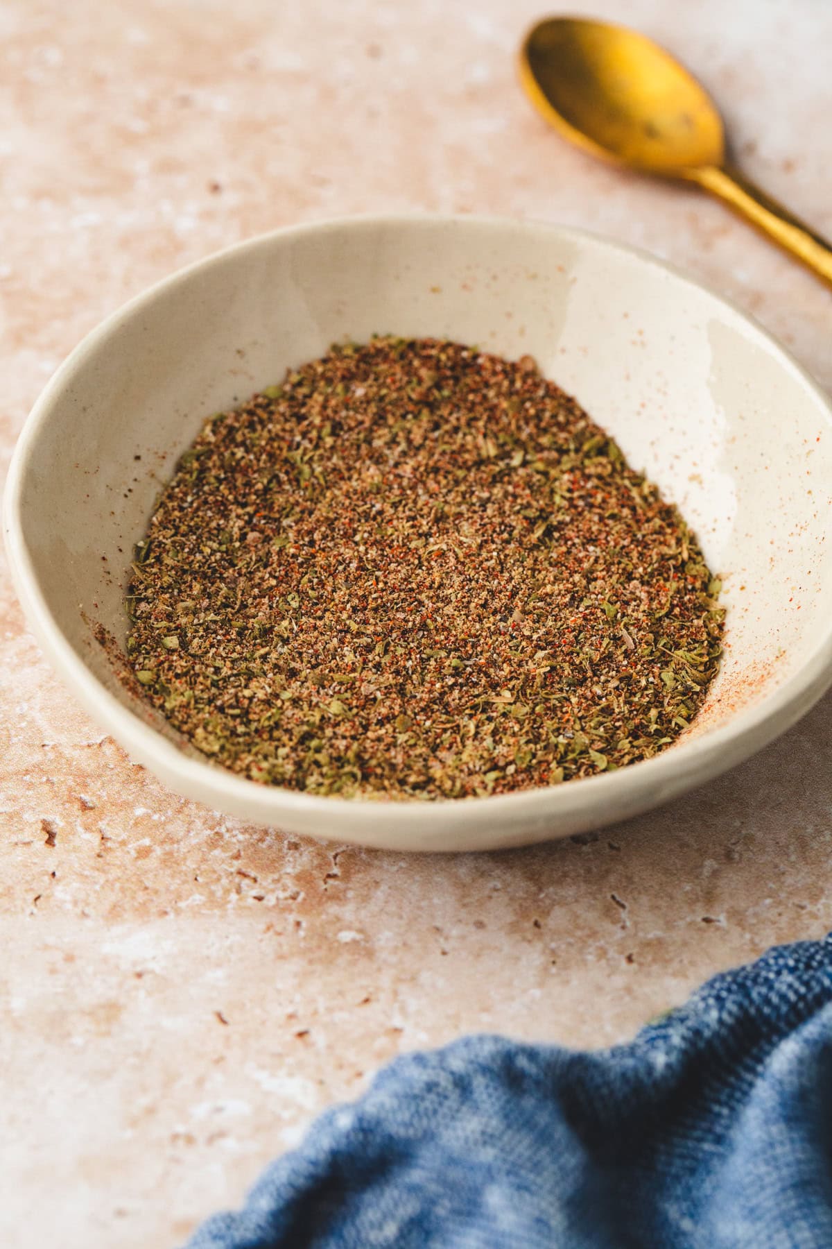 A white bowl filled with a mixed spice blend featuring an ancho rub sits on a beige textured surface. A golden spoon is placed nearby, with part of a blue cloth peeking from the corner.