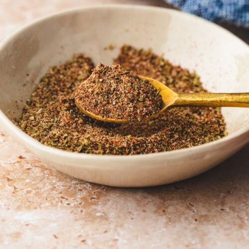 A beige bowl holds a coarse, multicolored spice blend, enhanced with ancho rub. A gold spoon rests gracefully in the bowl, slightly elevated, cradling the flavorful mix. The background showcases a blue textured fabric.
