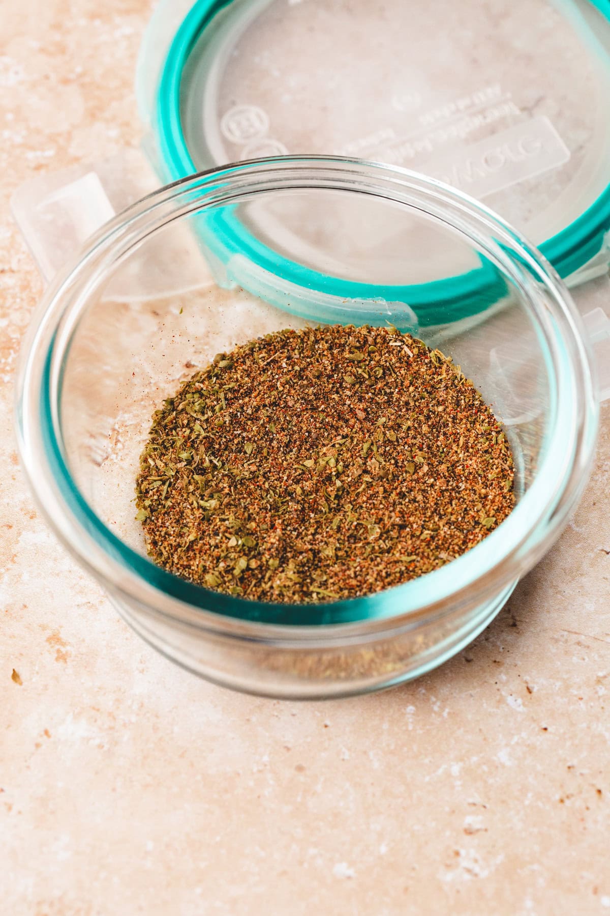 A clear glass bowl filled with a mix of ancho rub, ground spices, and herbs sits on a beige surface. The bowl is partially covered by a round, transparent lid with a teal rubber rim.