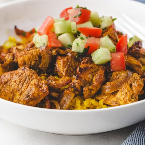 A bowl of spiced grilled meat on a bed of yellow rice, topped with diced cucumbers and tomatoes. A fork and a blue cloth are partially visible on the side.
