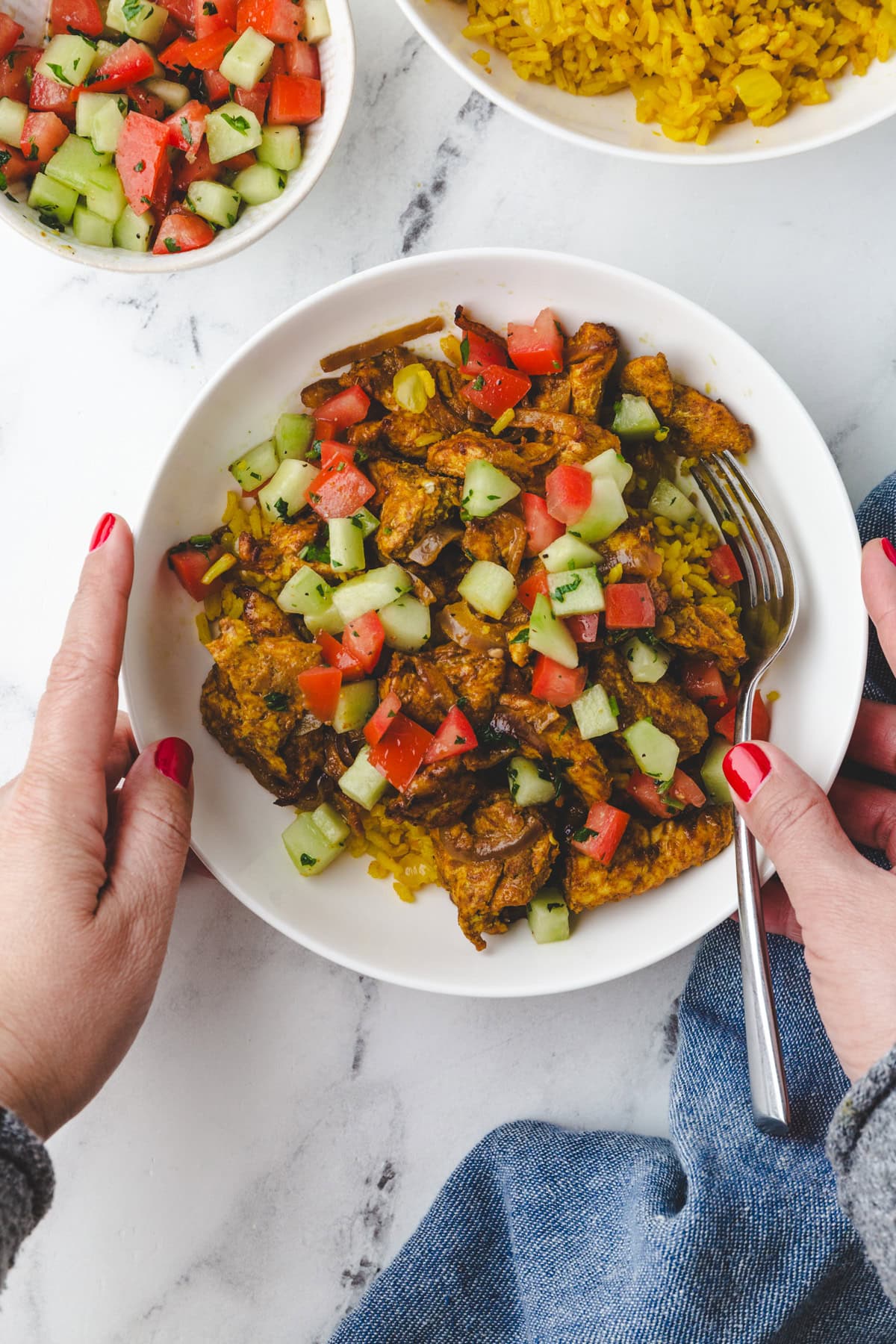 A person with red nail polish holds a bowl of food containing a chicken shawarma bowl , and a fresh salad of diced tomatoes and cucumbers. A fork is placed inside the bowl. A side of yellow rice and salad are partially visible nearby.