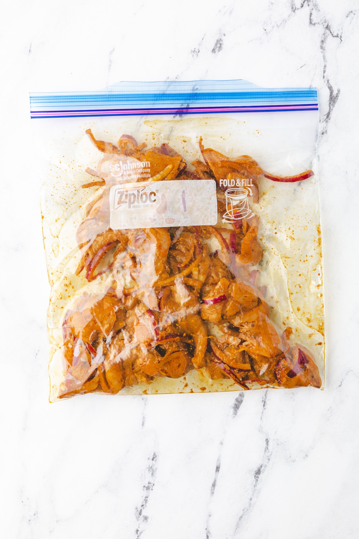 A Ziploc bag filled with marinated chicken slices mixed with red onion pieces. The bag is placed on a white marble surface, showcasing the spices and ingredients inside.