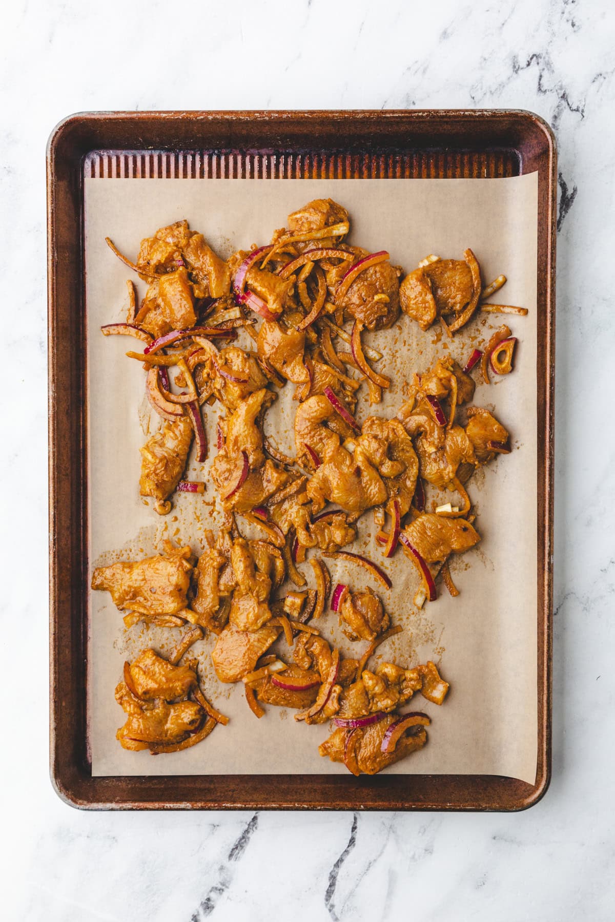 Pieces of seasoned chicken shawarma and sliced red onions spread out on parchment paper on a baking sheet, placed on a marble surface.