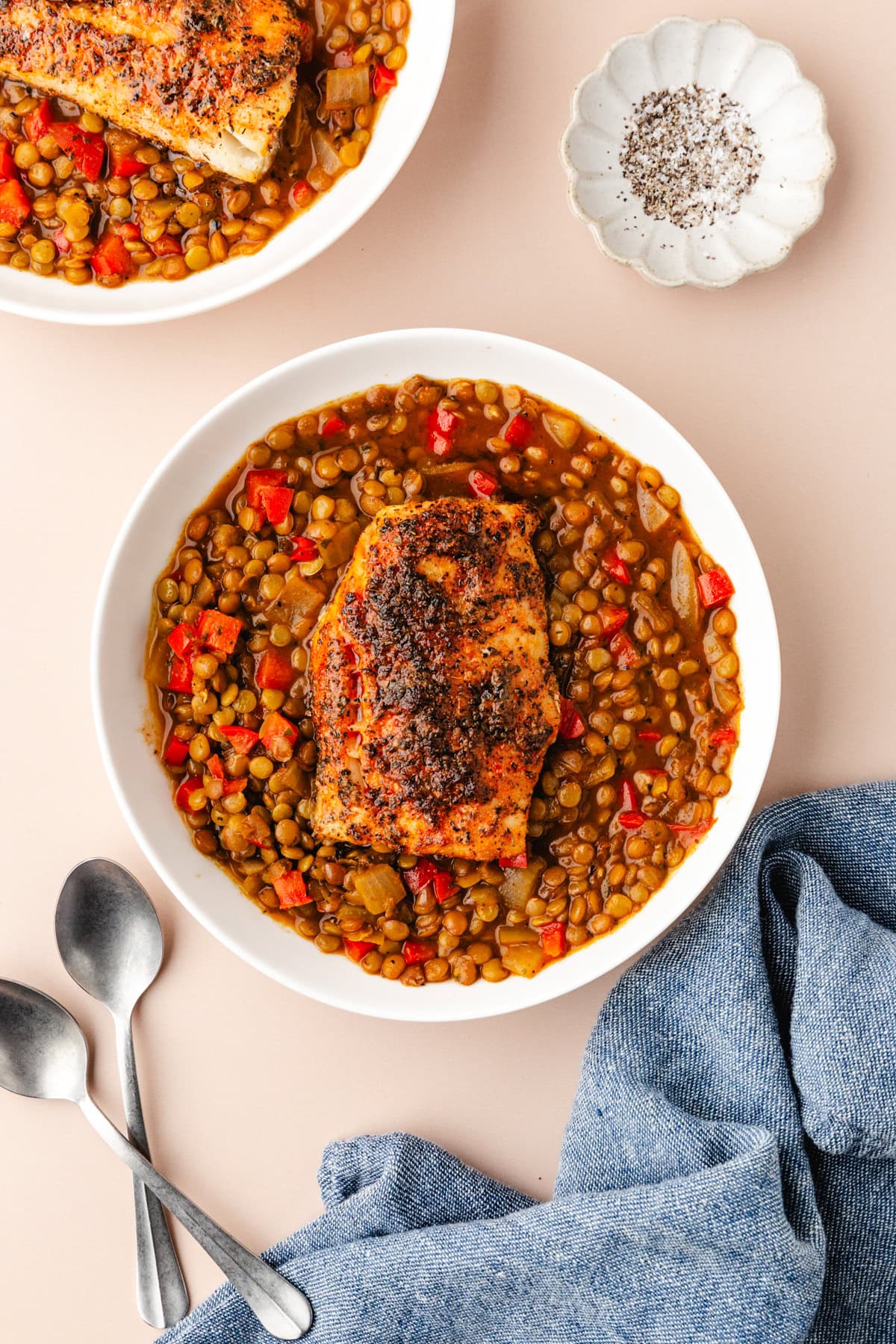 Two bowls of spiced roasted chicken on a bed of lentils with red peppers and onions. A small dish of pepper is nearby, along with two spoons and a blue napkin on a light surface.