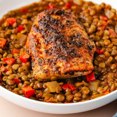 A white bowl filled with spiced lentils and diced red bell peppers, topped with a seasoned fillet of salmon. The dish sits on a light-colored surface.