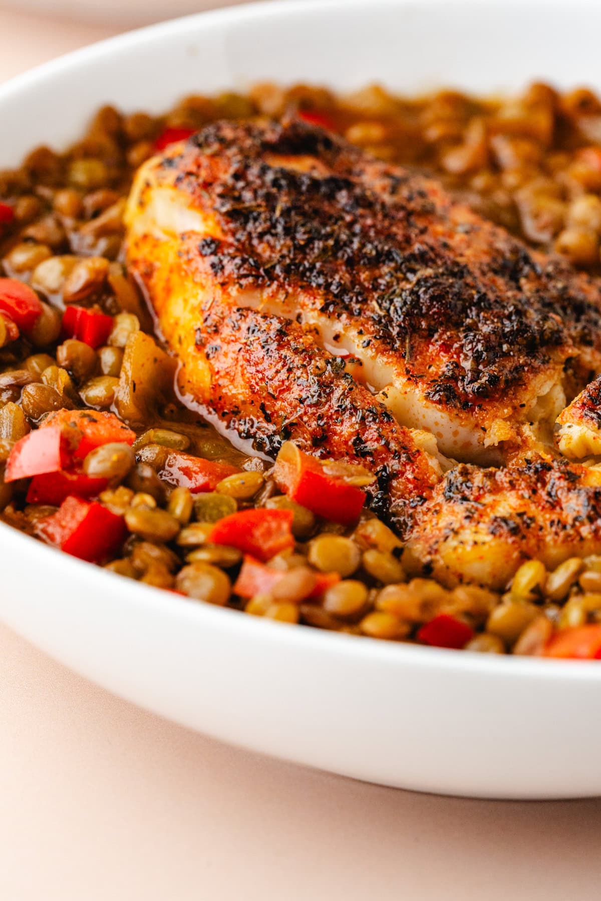 Close-up of a white bowl filled with seasoned, grilled chicken breast resting on a bed of lentils and diced red peppers. The dish is seasoned with visible spices, giving the chicken a crispy, flavorful texture.