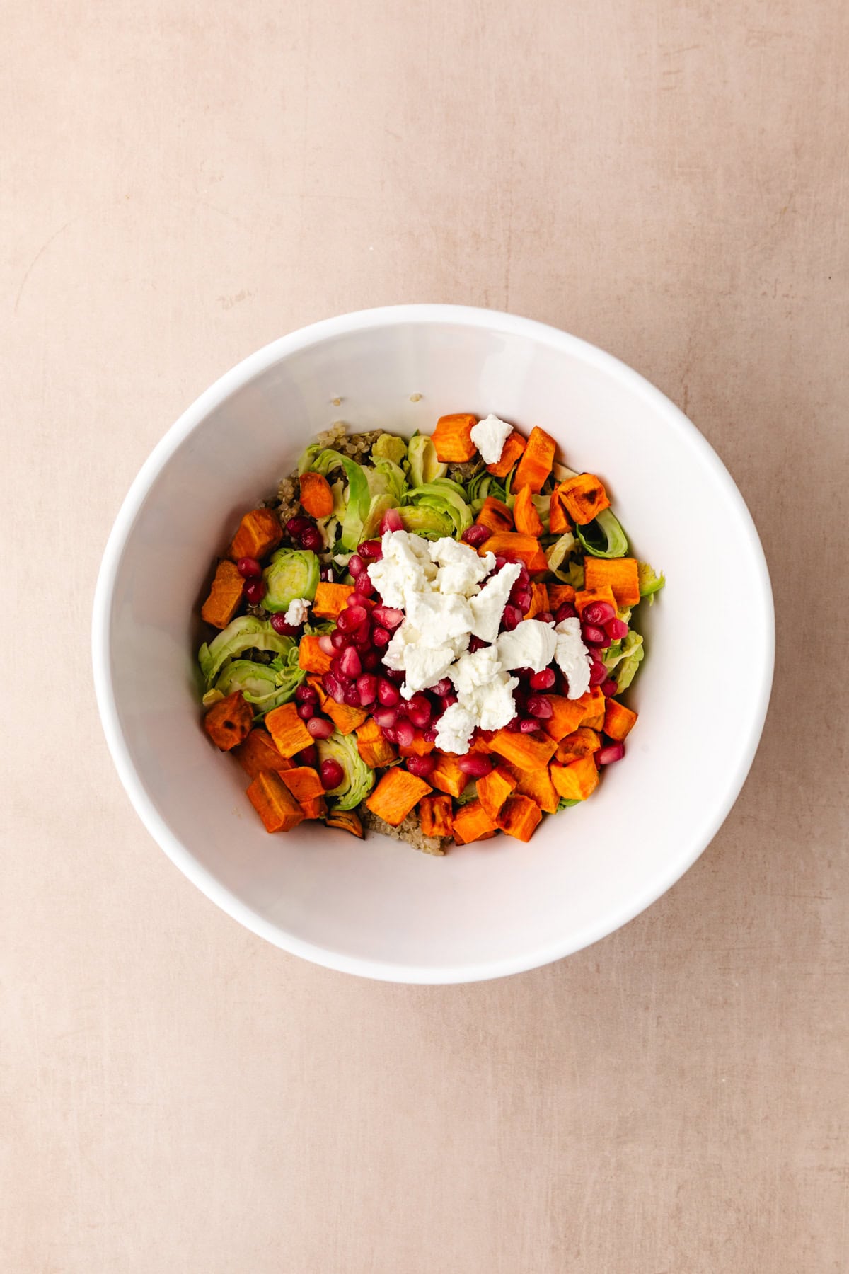 A white bowl filled with a colorful salad, including quinoa, roasted sweet potatoes, Brussels sprouts, pomegranate seeds, and crumbled feta cheese. The dish is set against a light beige background.
