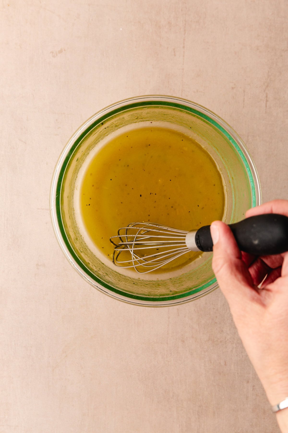 A person is whisking a yellowish-green liquid in a clear glass bowl on a beige countertop. The liquid appears to be a dressing or sauce. The whisk has a black handle, and the bowl is viewed from above.