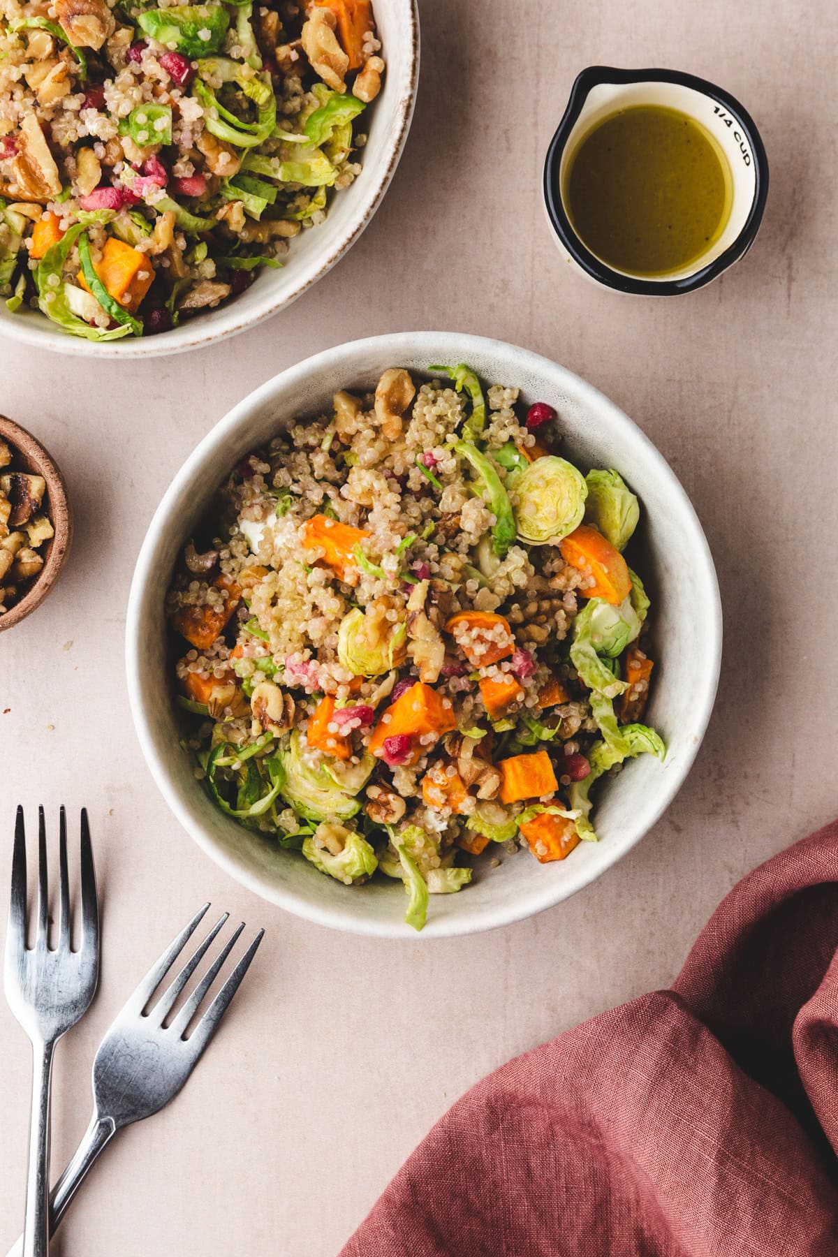 A bowl of quinoa salad with roasted sweet potatoes, Brussels sprouts, walnuts, and pomegranate seeds. A small dish of vinaigrette is beside the bowl. Two forks and a maroon napkin are also on the table.