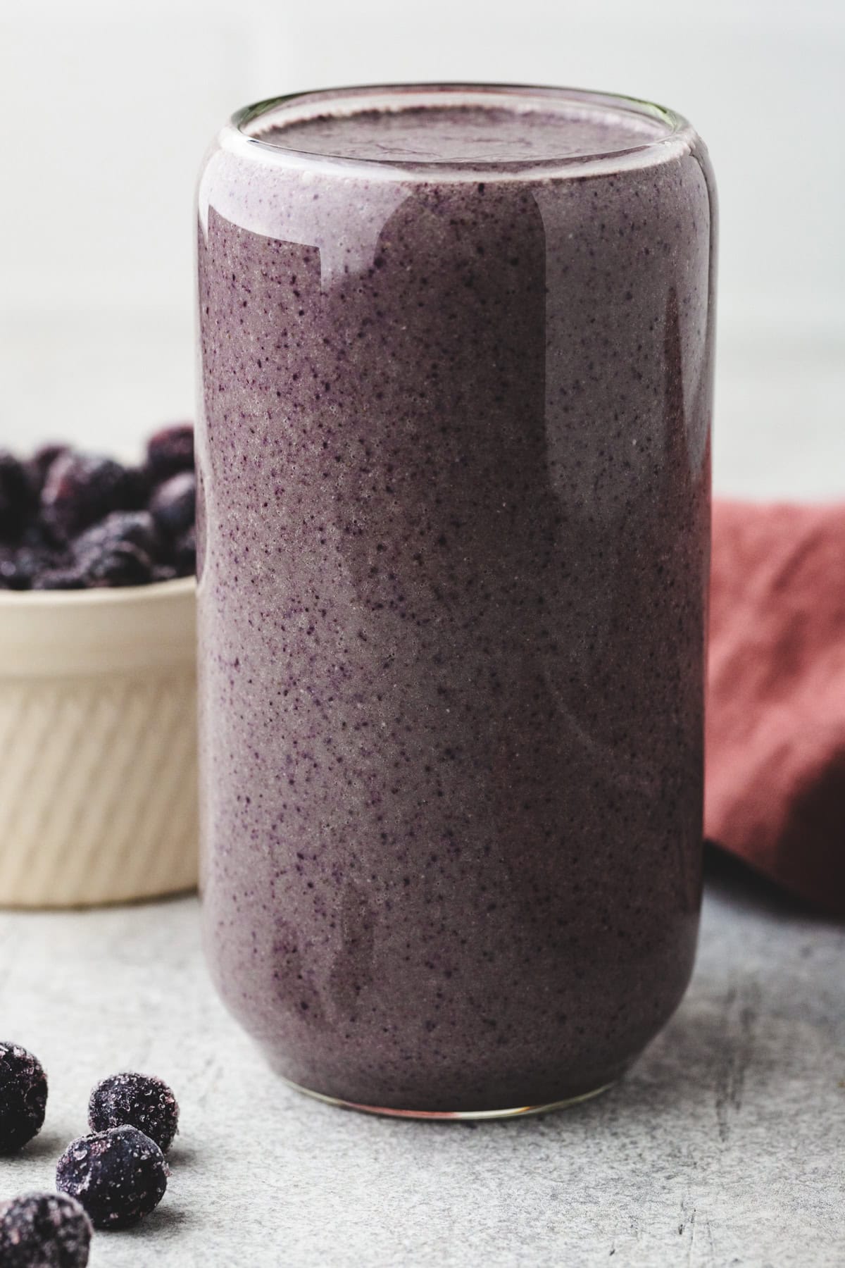 A tall glass filled with a thick, purple smoothie is placed on a countertop. Beside it are a few blackberries and a bowl containing more berries. A red cloth napkin is partially visible in the background.