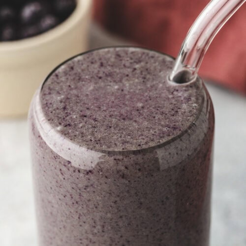 A tall glass filled with a thick purple smoothie, with a reusable clear straw inserted. In the background, theres a bowl of blueberries and a soft-focus reddish cloth.