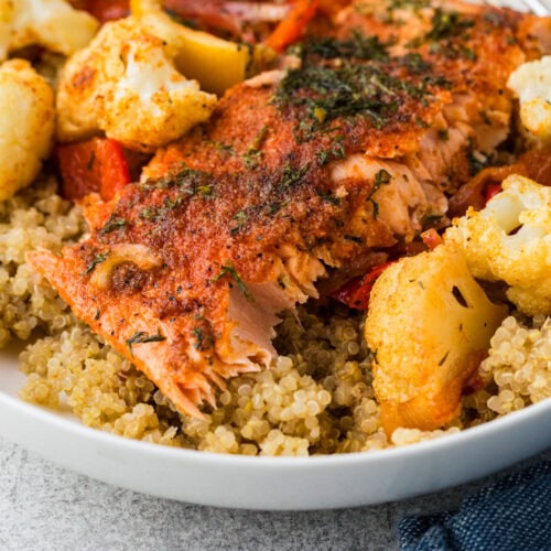 A close-up of a dish featuring a seasoned, cooked salmon fillet atop a bed of quinoa. Surrounding the salmon are roasted cauliflower florets, red bell peppers, and potato chunks. A hint of dill garnishes the meal, and a blue napkin is nearby.