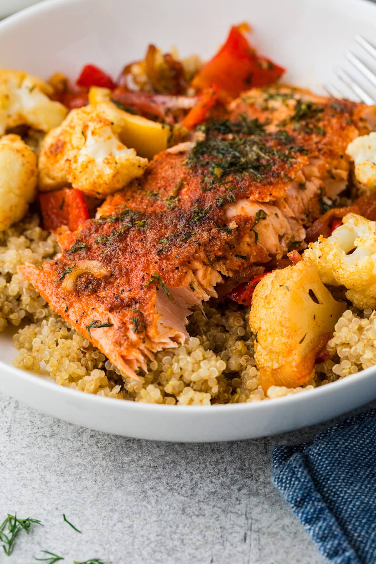 A close-up of a dish featuring a seasoned, cooked salmon fillet atop a bed of quinoa. Surrounding the salmon are roasted cauliflower florets, red bell peppers, and potato chunks. A hint of dill garnishes the meal, and a blue napkin is nearby.